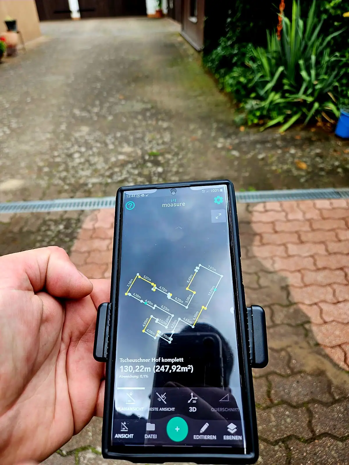 Mobilgerät in der Hand vor einer Pflasterfläche, auf dem Bildschirm der digital erfasste Grundriss mit Maßangaben.